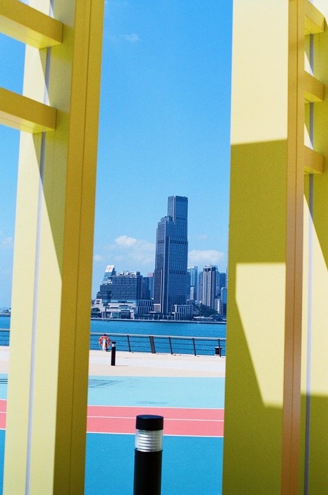 A tall building stands across a body of water, framed by yellow structures on either side.