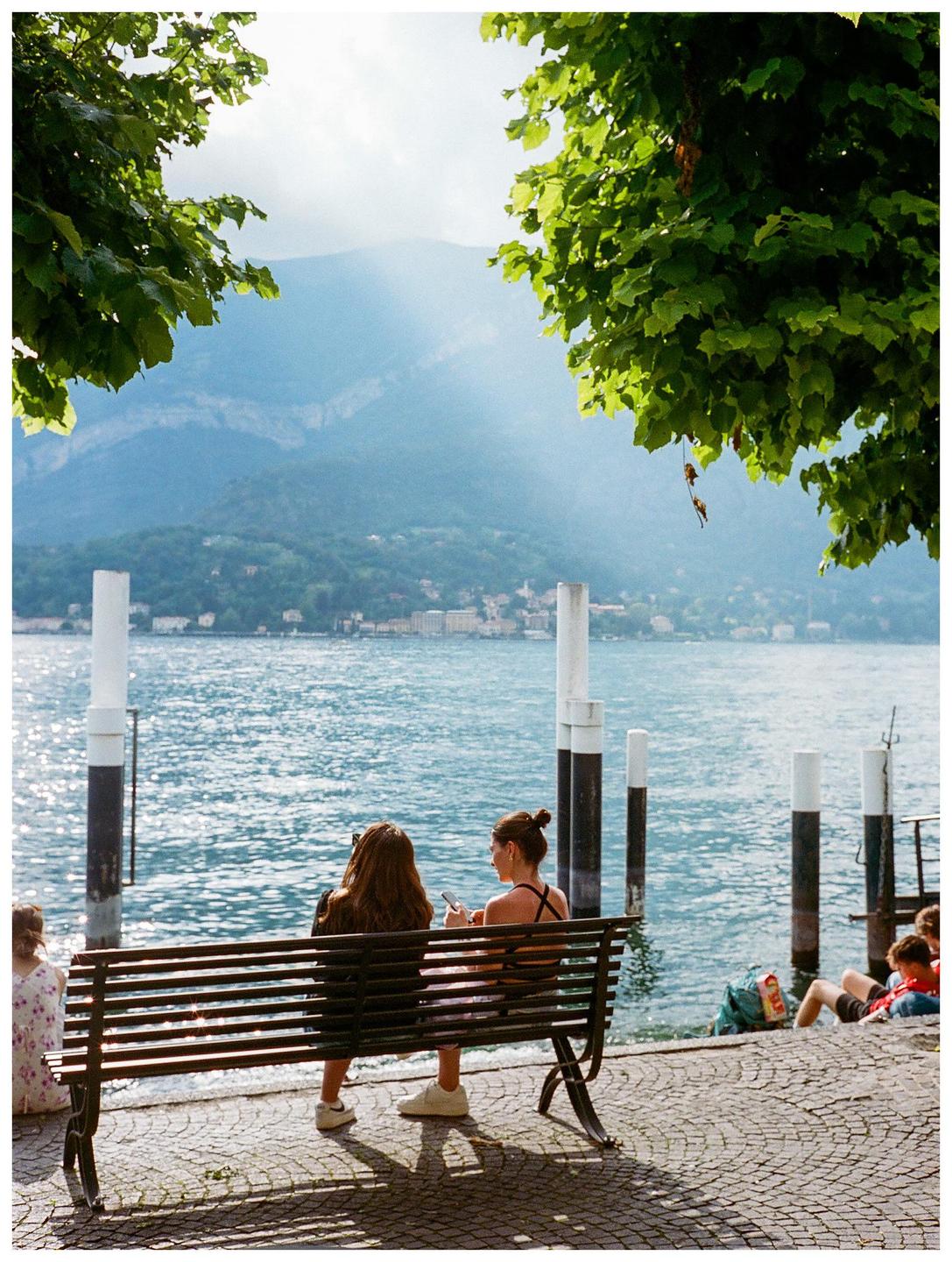 Two women sit on a bench by a lake with mountains in the background.