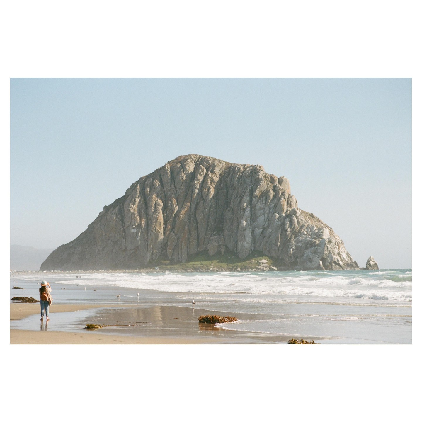 A woman stands on a beach looking at a large rock formation in the ocean.