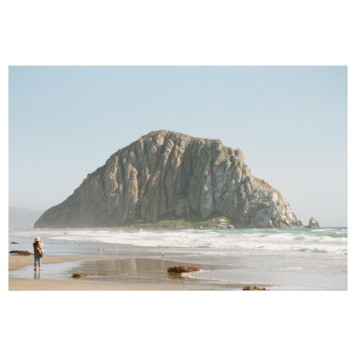 A woman stands on a beach looking at a large rock formation in the ocean.
