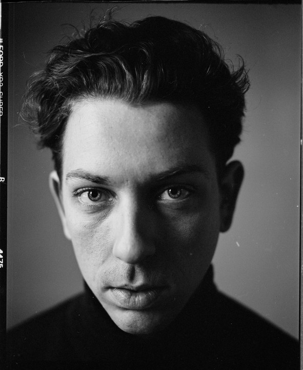 A man with curly hair and a dark turtleneck stares directly at the viewer.
