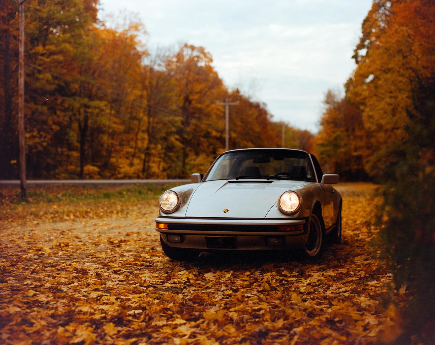 A silver car with its headlights on is parked on a road covered in autumn leaves, surrounded by trees with orange foliage.