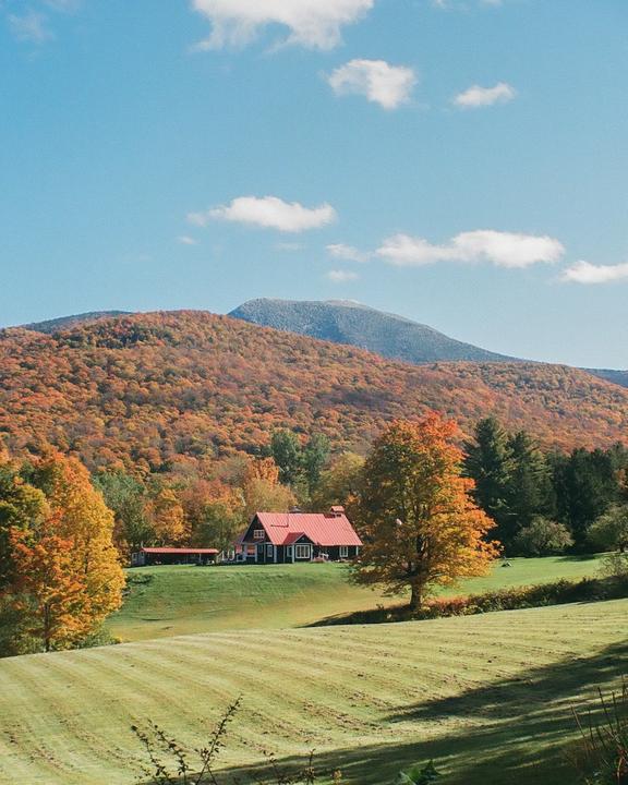 A house with a red roof sits on a green hill with a forest of autumn trees and a mountain in the background.