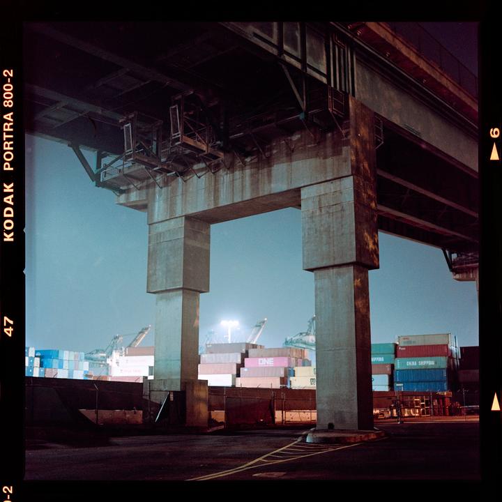 A concrete overpass looms over a cargo port at night, with stacks of shipping containers visible in the distance.