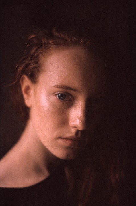 A woman with red hair and blue eyes looks directly at the viewer, with half of her face in shadow.