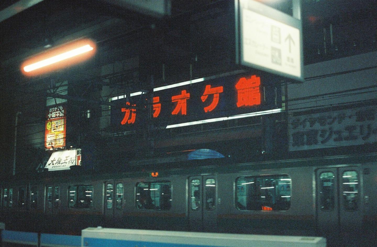 A train is stopped at a station platform at night, with neon signs visible above it.