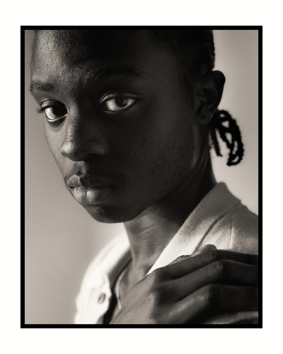 A man with braided hair looks directly at the viewer while holding his shirt collar.