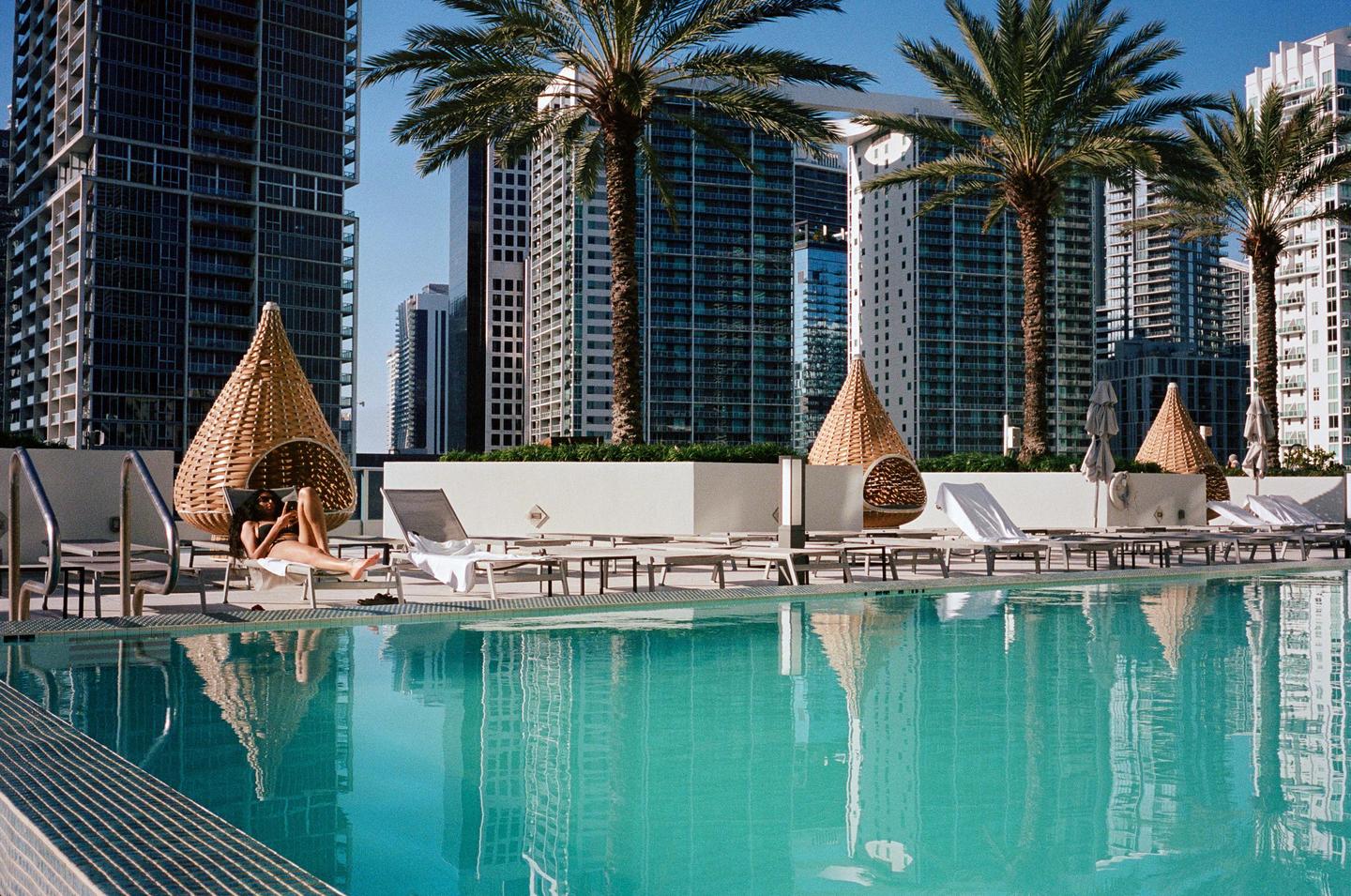 A woman relaxes on a lounge chair by a swimming pool surrounded by tall buildings and palm trees.