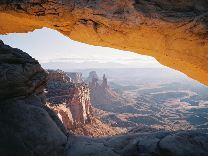 A natural rock arch frames a vast desert landscape with distant mountains under a bright sky.