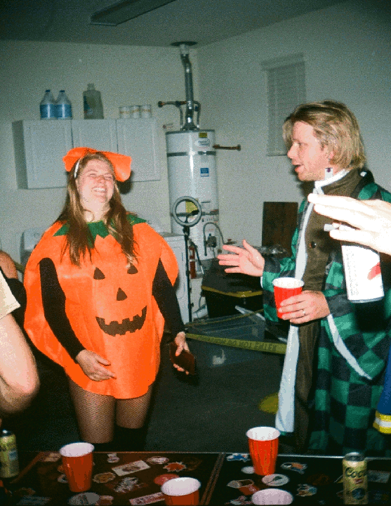 A woman in a pumpkin costume laughs while a man in a green and black checkered robe talks to her at a party.