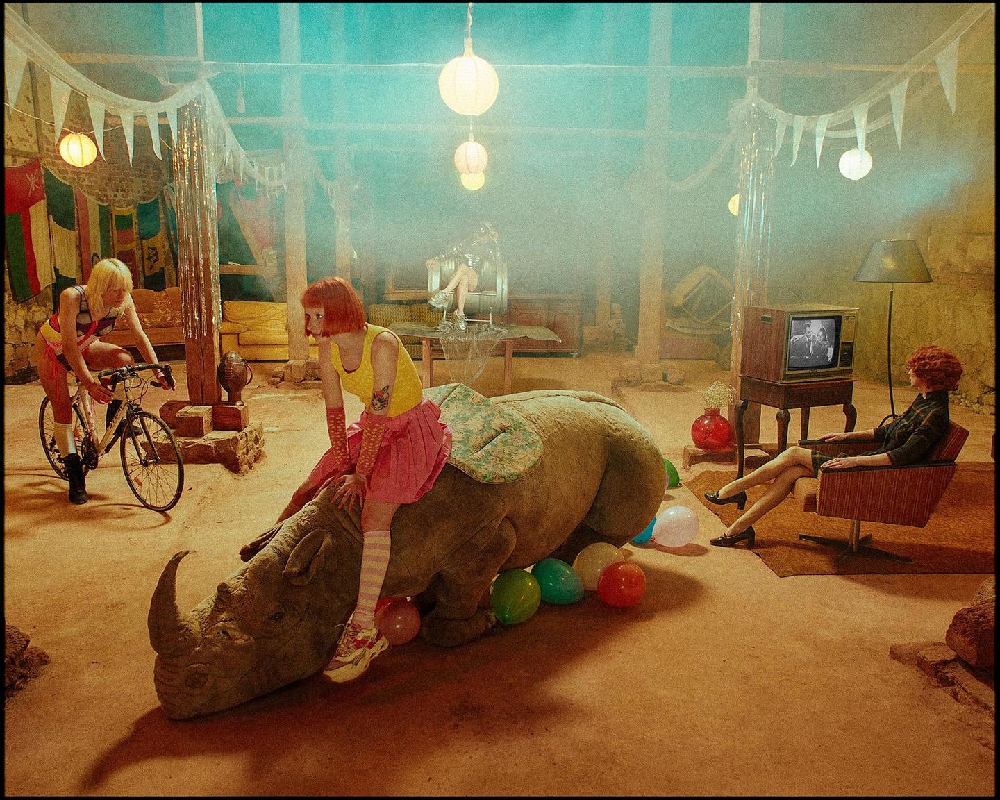 Three women are in a hazy room with a rhinoceros, balloons, and a television.