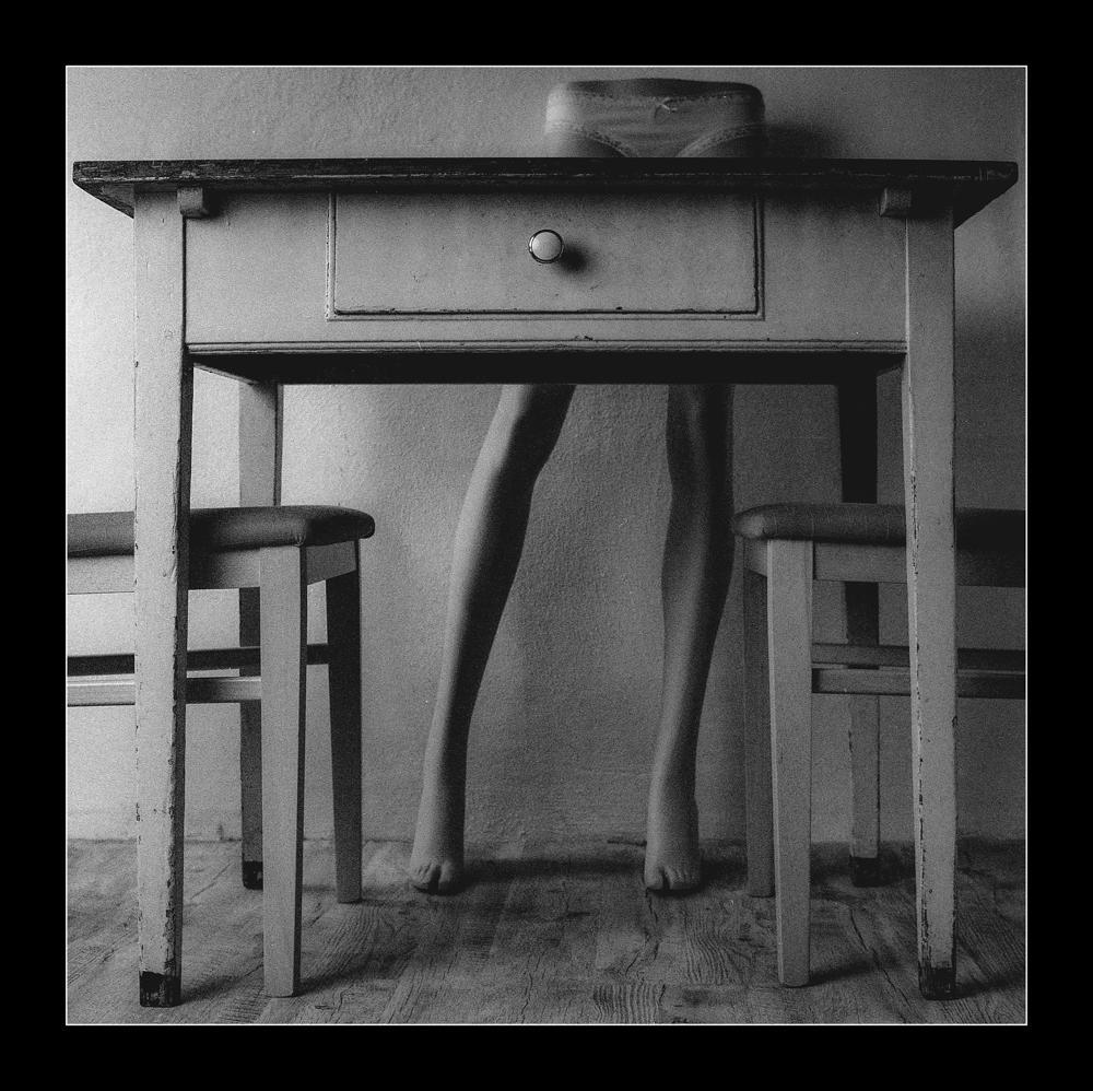 A person's legs are visible under a table with two chairs and underwear on top.