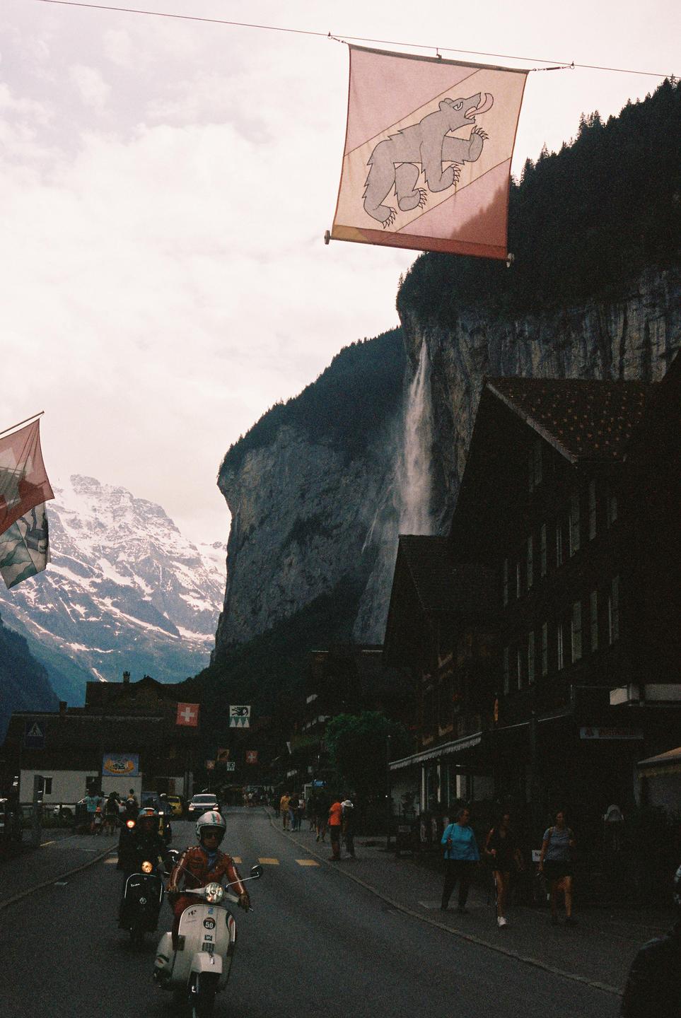 A street in Lauterbrunnen is filled with people and scooters, with a waterfall cascading down a cliff face and snow-capped mountains in the background.