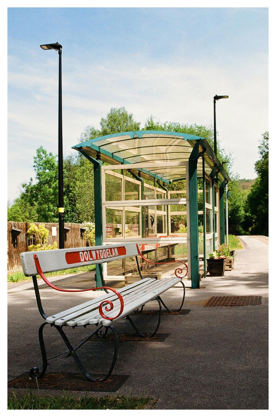 A white and red bench sits in front of a glass bus stop shelter on a sunny day.