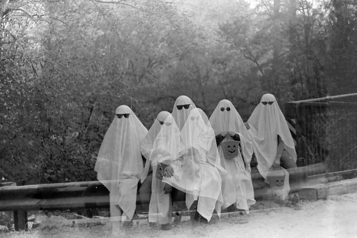 Six people dressed as ghosts with sunglasses sit on a guardrail, with one holding a jack-o'-lantern bucket.