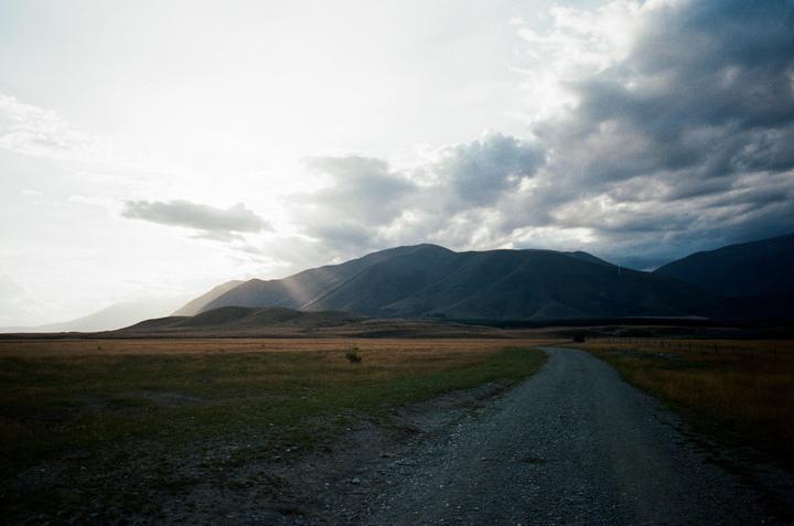 A gravel road leads through a field towards mountains under a cloudy sky with sun rays breaking through.