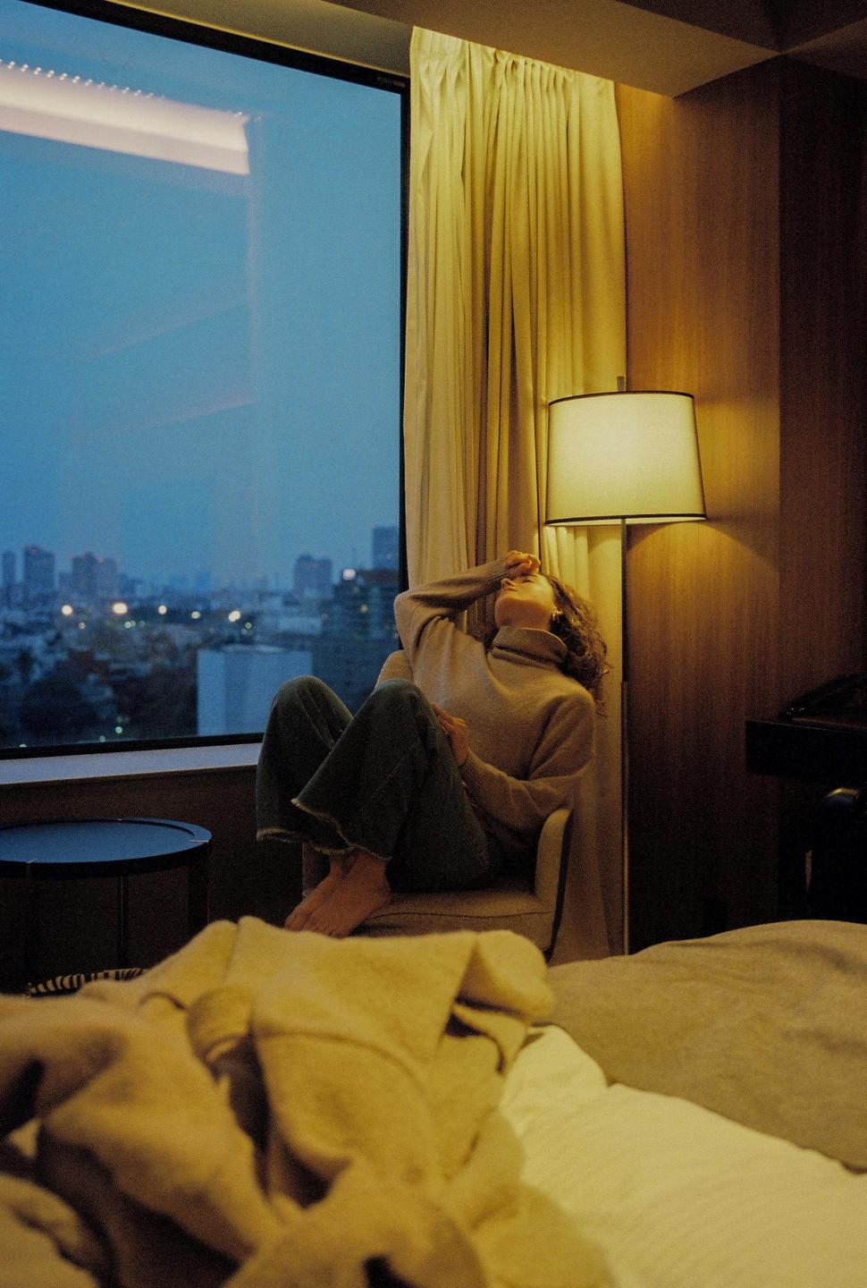 A woman sits in a chair next to a window overlooking a city at dusk.