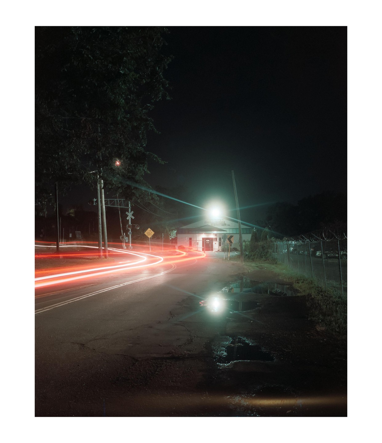 Red light trails from a vehicle illuminate a wet road at night, with a building and railroad crossing in the background.