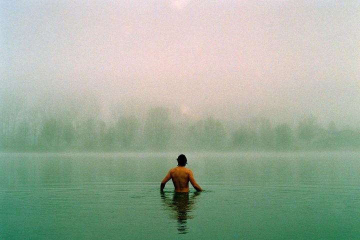 A man stands in a body of water with a hazy, tree-lined shore in the distance.