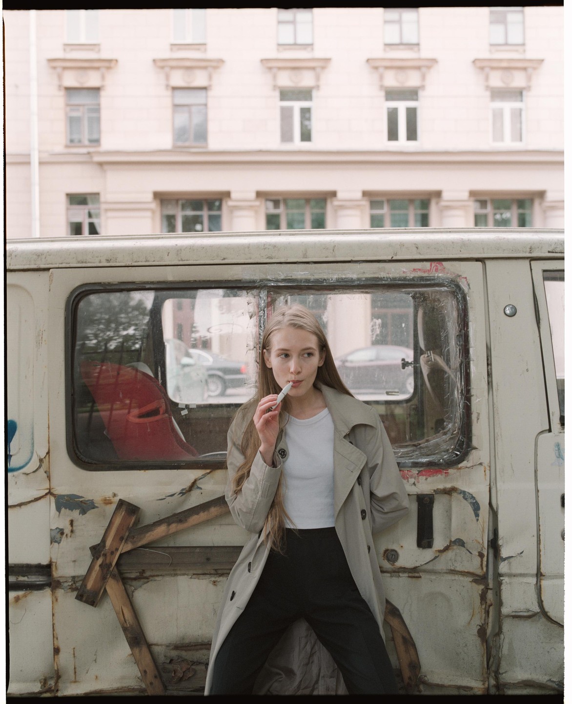 A woman in a trench coat stands next to a weathered van on a street.