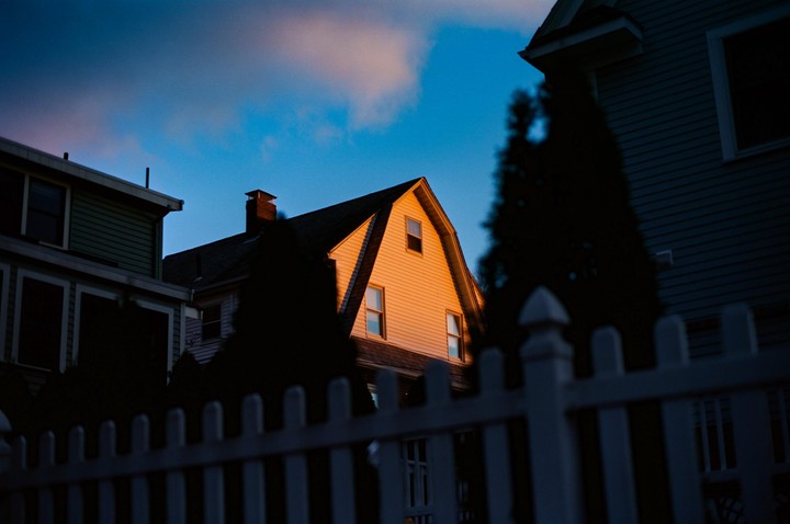 The side of a house is illuminated by golden light against a deep blue sky, framed by a white picket fence and dark trees.