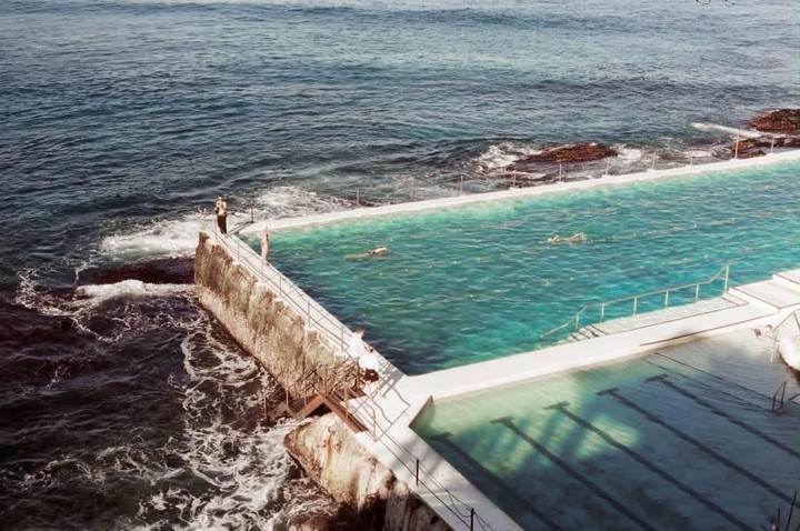 A swimming pool built into the ocean is filled with swimmers and surrounded by crashing waves.