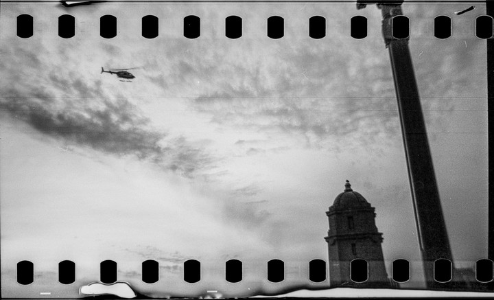 A helicopter flies over a domed tower and a tall column against a cloudy sky.