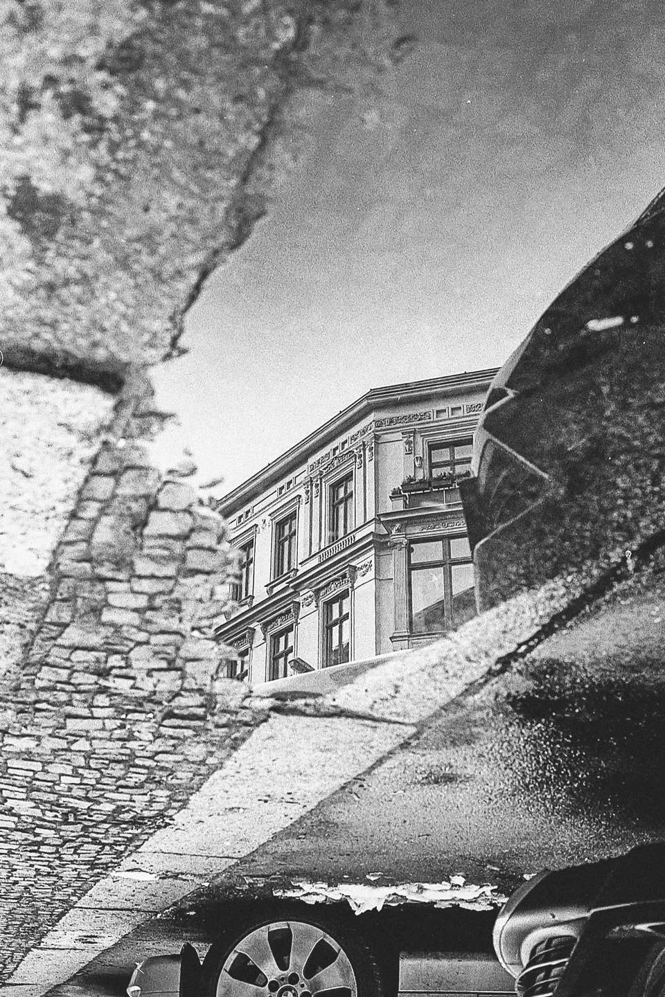 A building and a car are reflected upside down in a puddle on a cobblestone street.