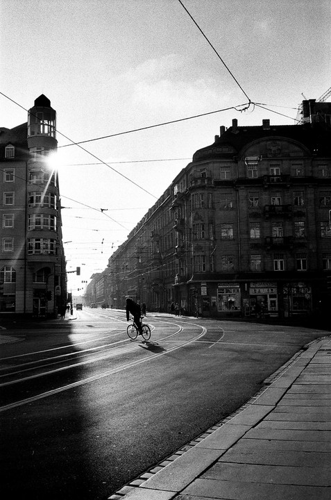 A person rides a bicycle on a city street with buildings and overhead wires under a bright sky.