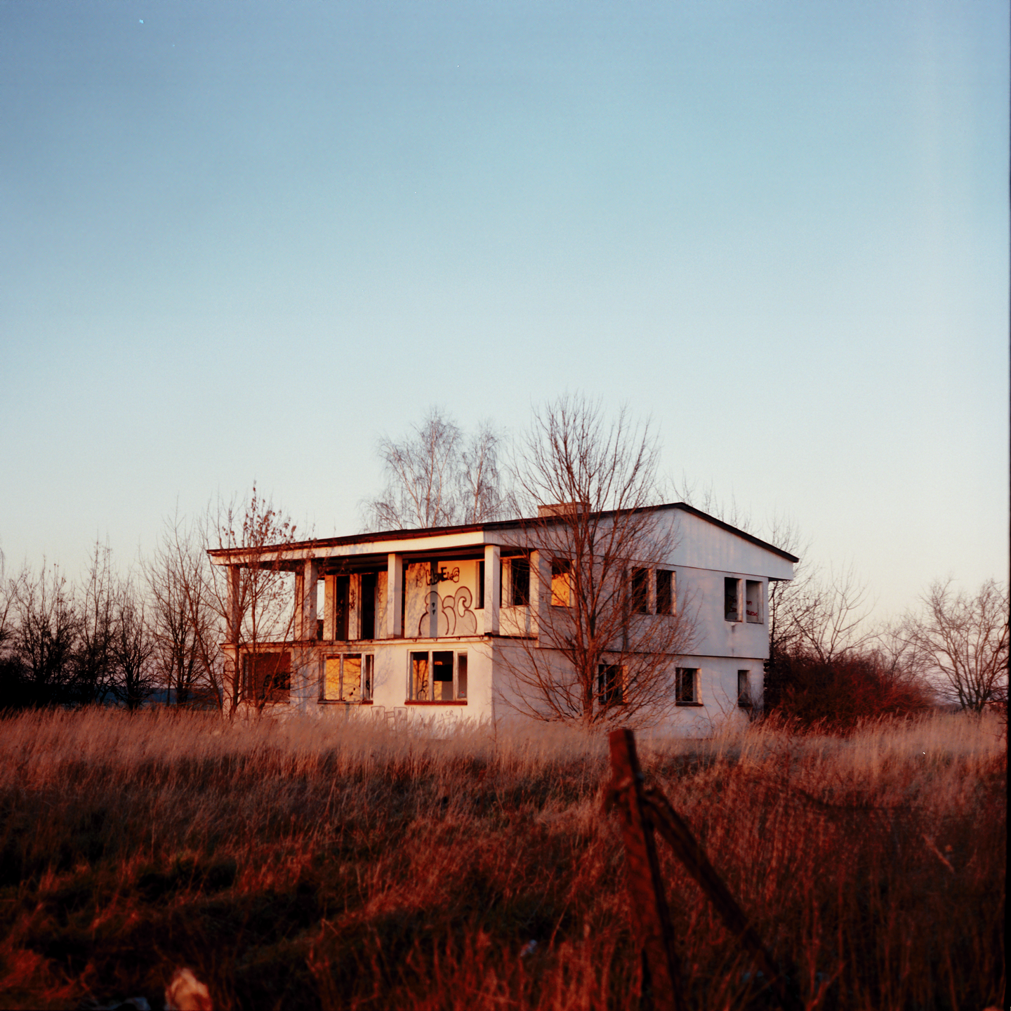 An abandoned two-story building with graffiti on its facade stands in a field of tall, dry grass under a clear sky.