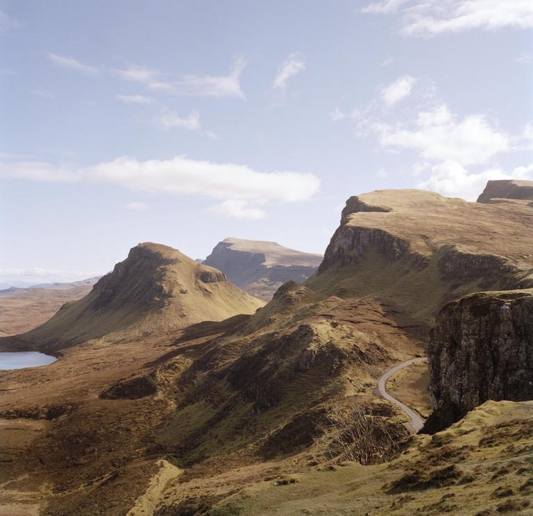 A winding road cuts through a vast, mountainous landscape with a body of water visible in the distance.