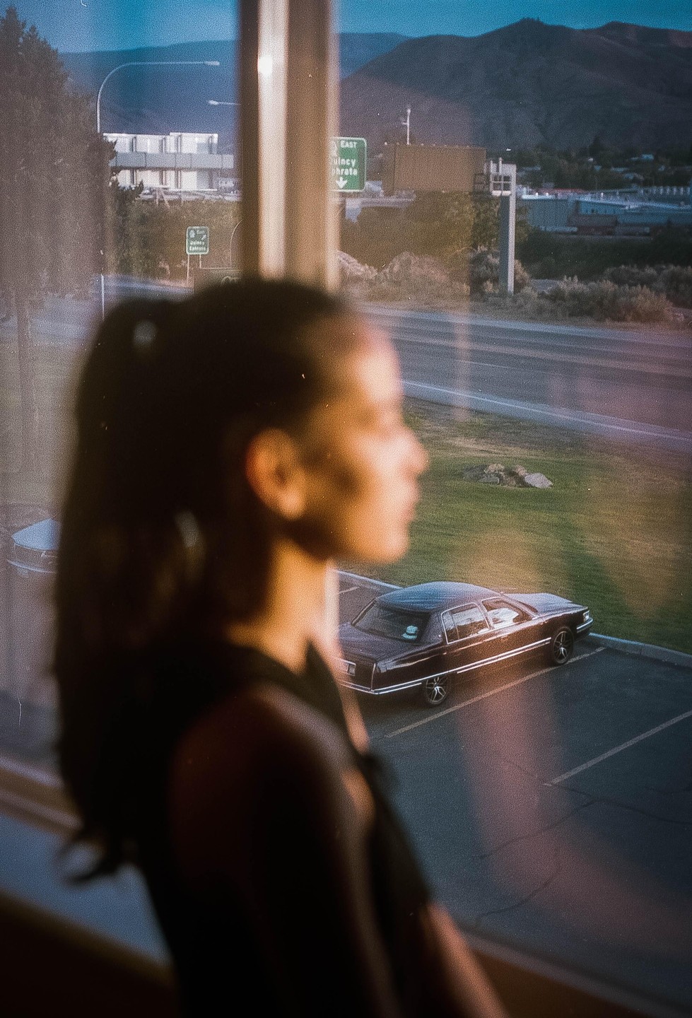 A woman looks out a window at a parking lot with a car and a highway sign for Quincy and Ephrata.