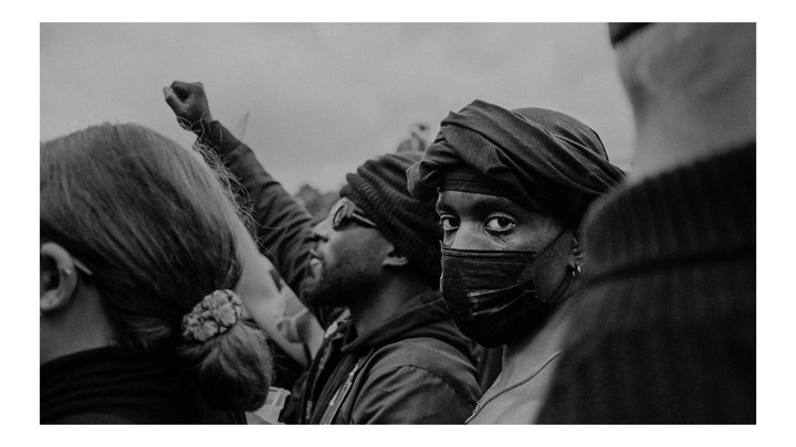 A person wearing a face mask and head covering looks directly at the viewer from a crowd of people.