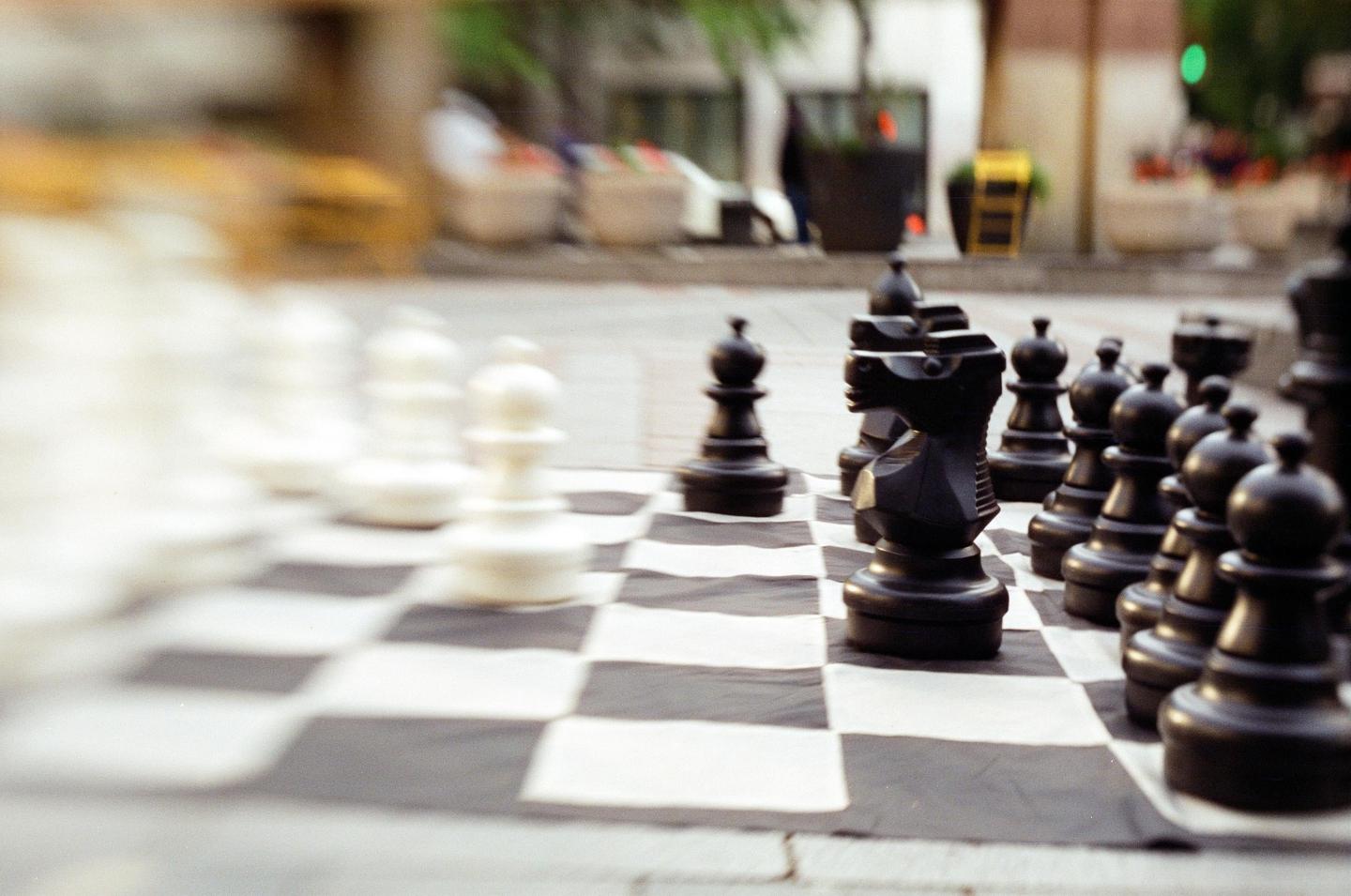 A large outdoor chessboard with black chess pieces in focus and white chess pieces blurred in the background.