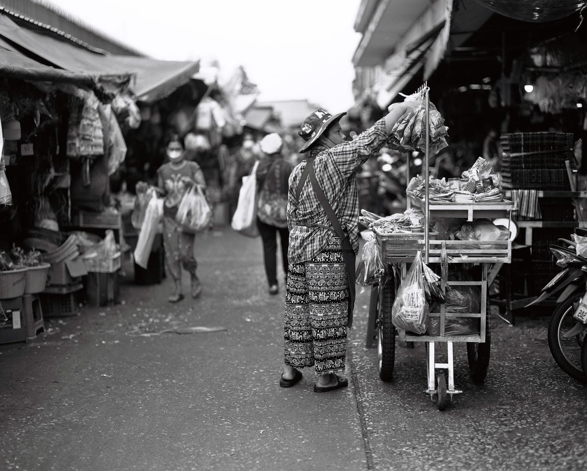 A woman wearing a hat and patterned pants is standing next to a cart filled with goods on a street.