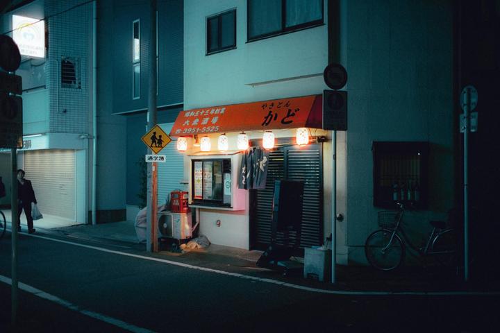 A person walks past a small shop with glowing lanterns and a bicycle parked outside at night.