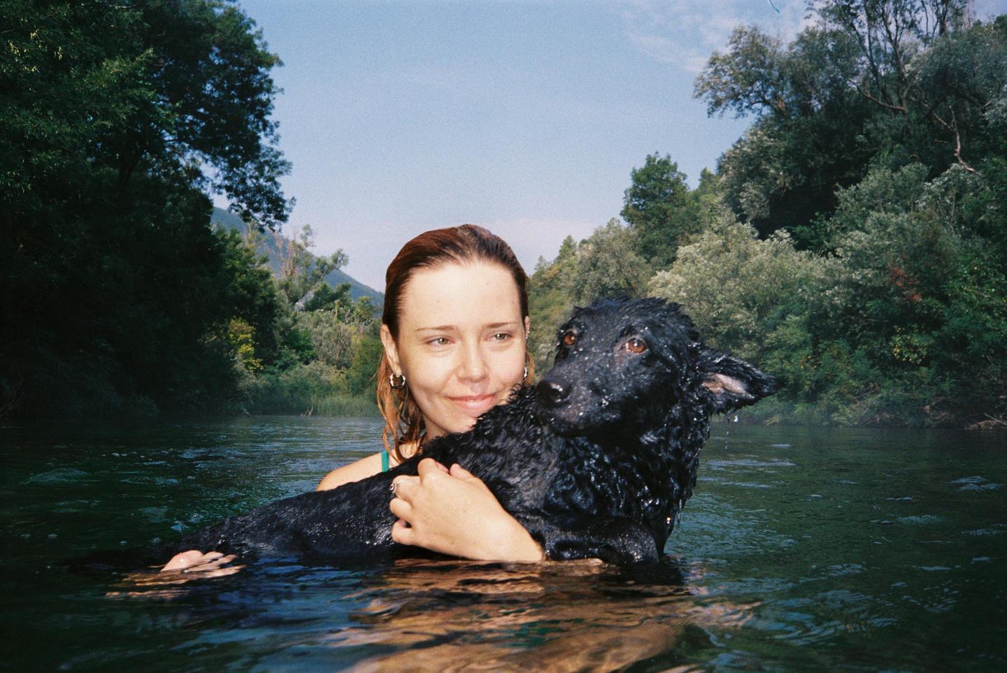 A woman holds a wet black dog in a river surrounded by lush green trees.