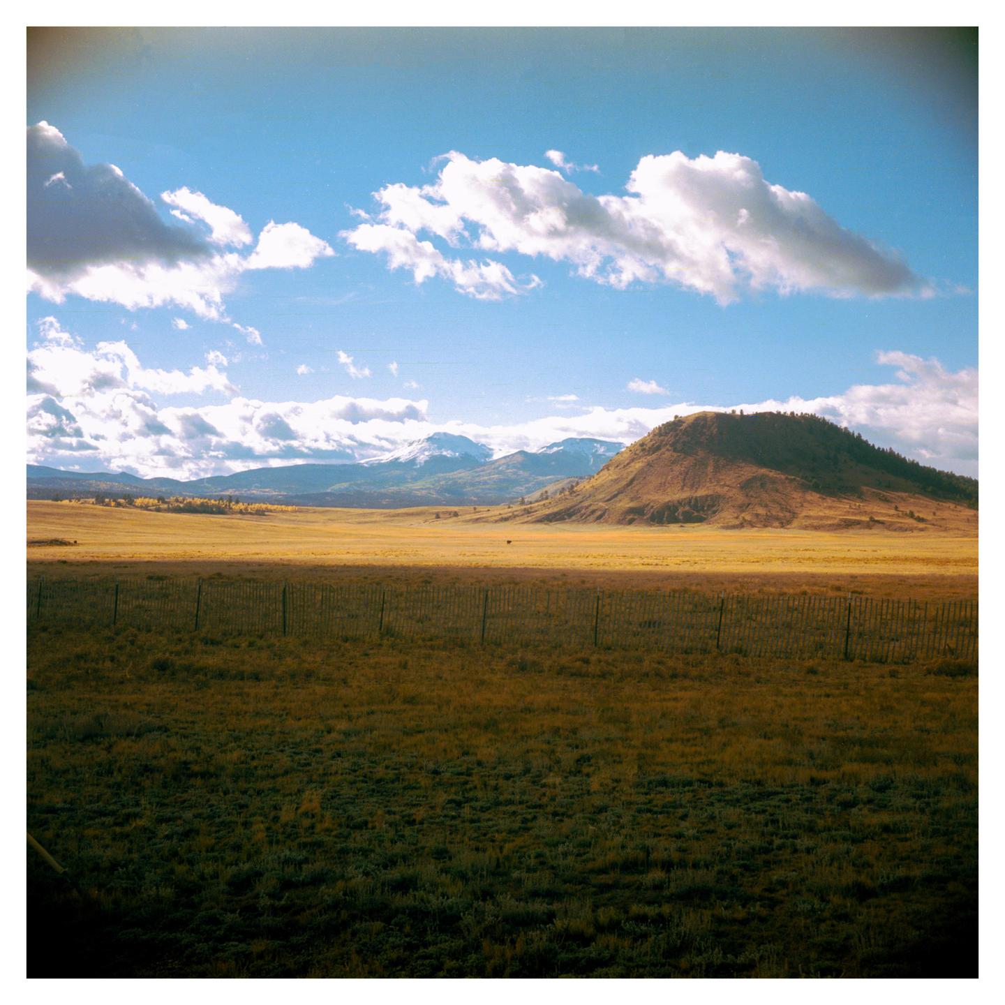 A fence separates a sunlit field from a shadowed field, with mountains and a hill in the background under a cloudy sky.