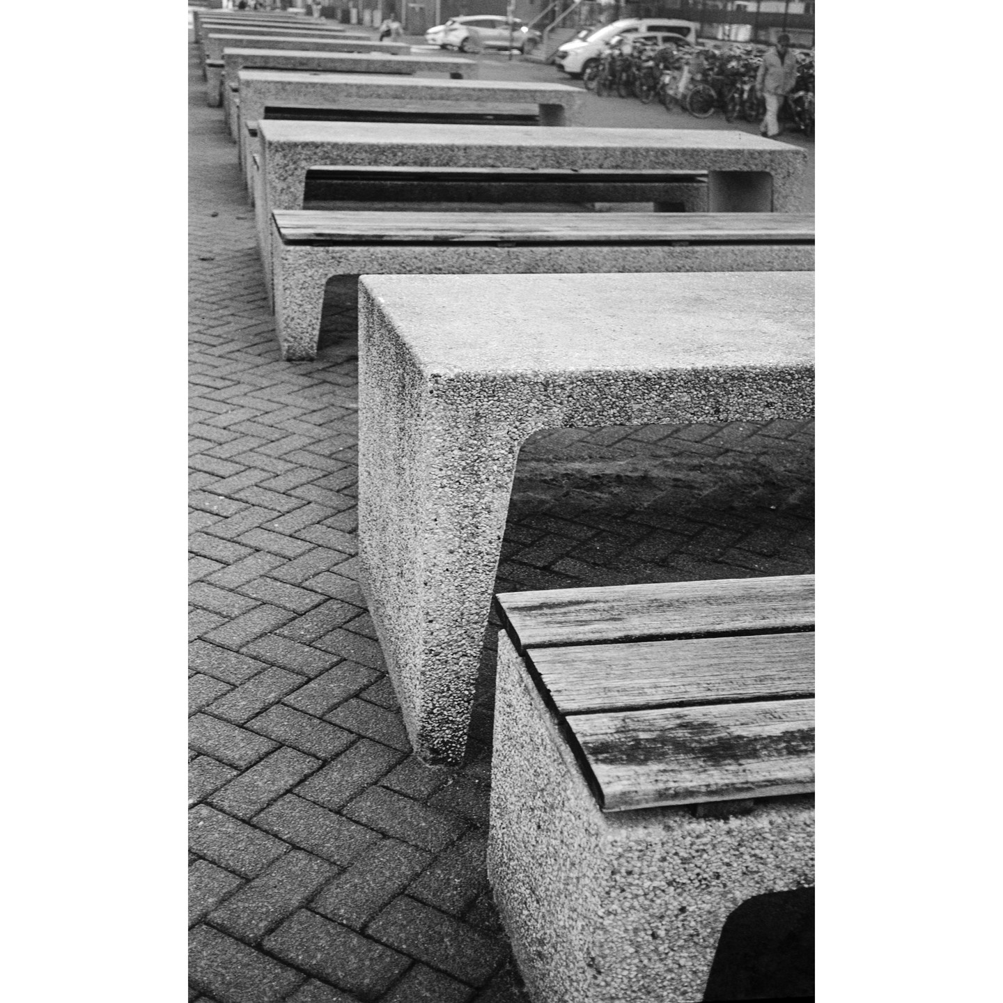 A row of concrete benches with wooden seats lines a brick sidewalk, with cars and bicycles visible in the background.
