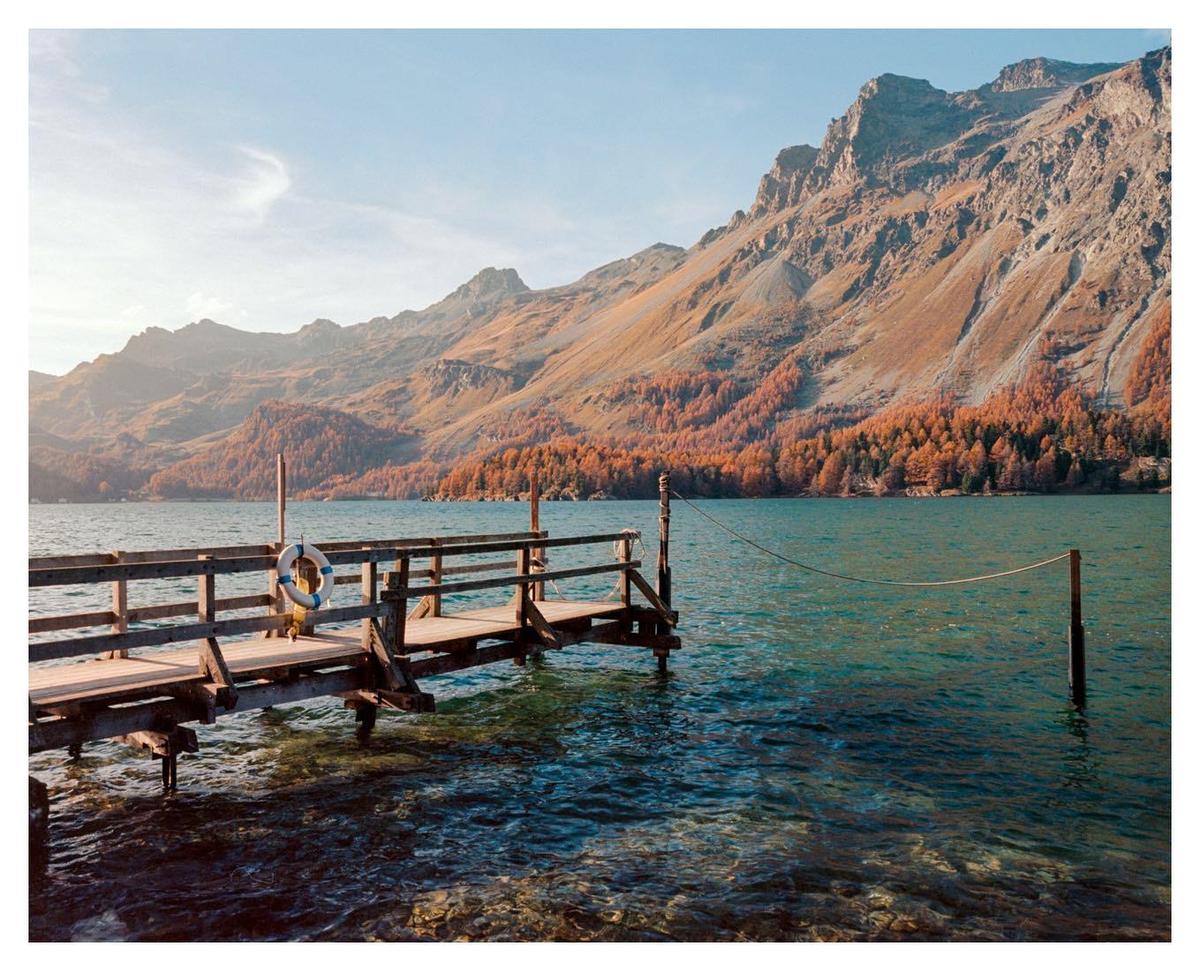 A wooden pier with a life preserver extends into a lake with mountains and autumn trees in the background.