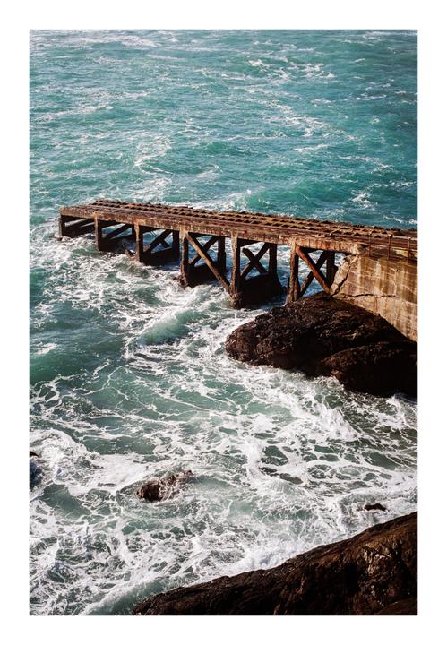 A weathered concrete pier extends into the turquoise ocean, with waves crashing against its supports and the rocky shore.