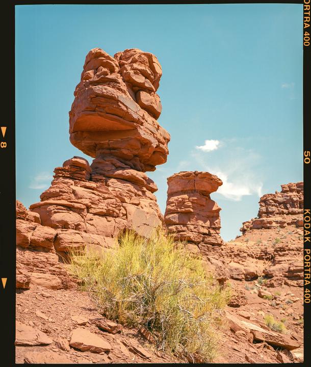 A large, balanced rock formation stands prominently against a clear blue sky, with smaller rock formations and desert brush in the foreground.
