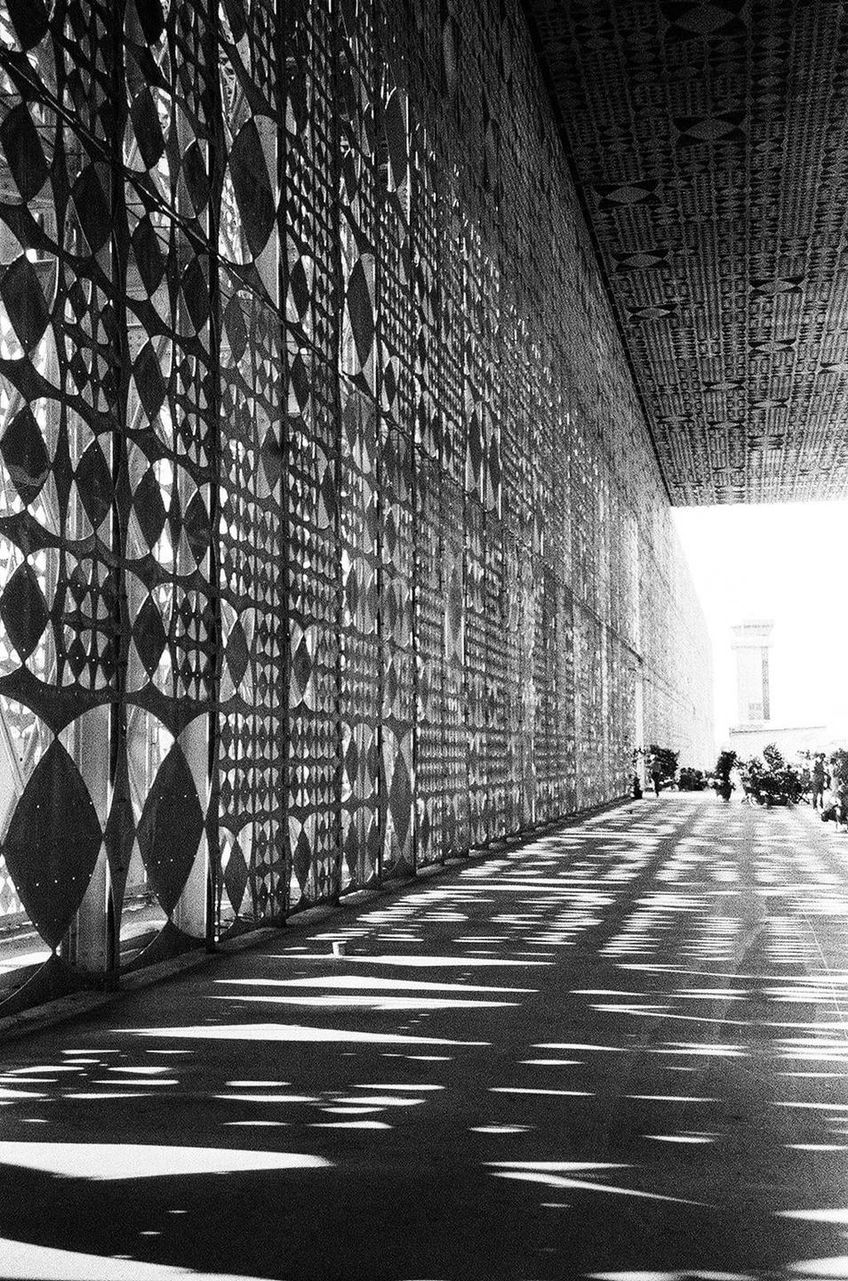 A long patterned wall casts shadows on the ground, leading towards a bright opening with distant buildings and people.