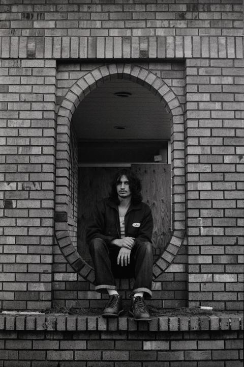 A man with long hair sits in an arched brick doorway.