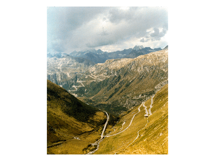 A winding road ascends a steep, grassy mountain valley under a cloudy sky.