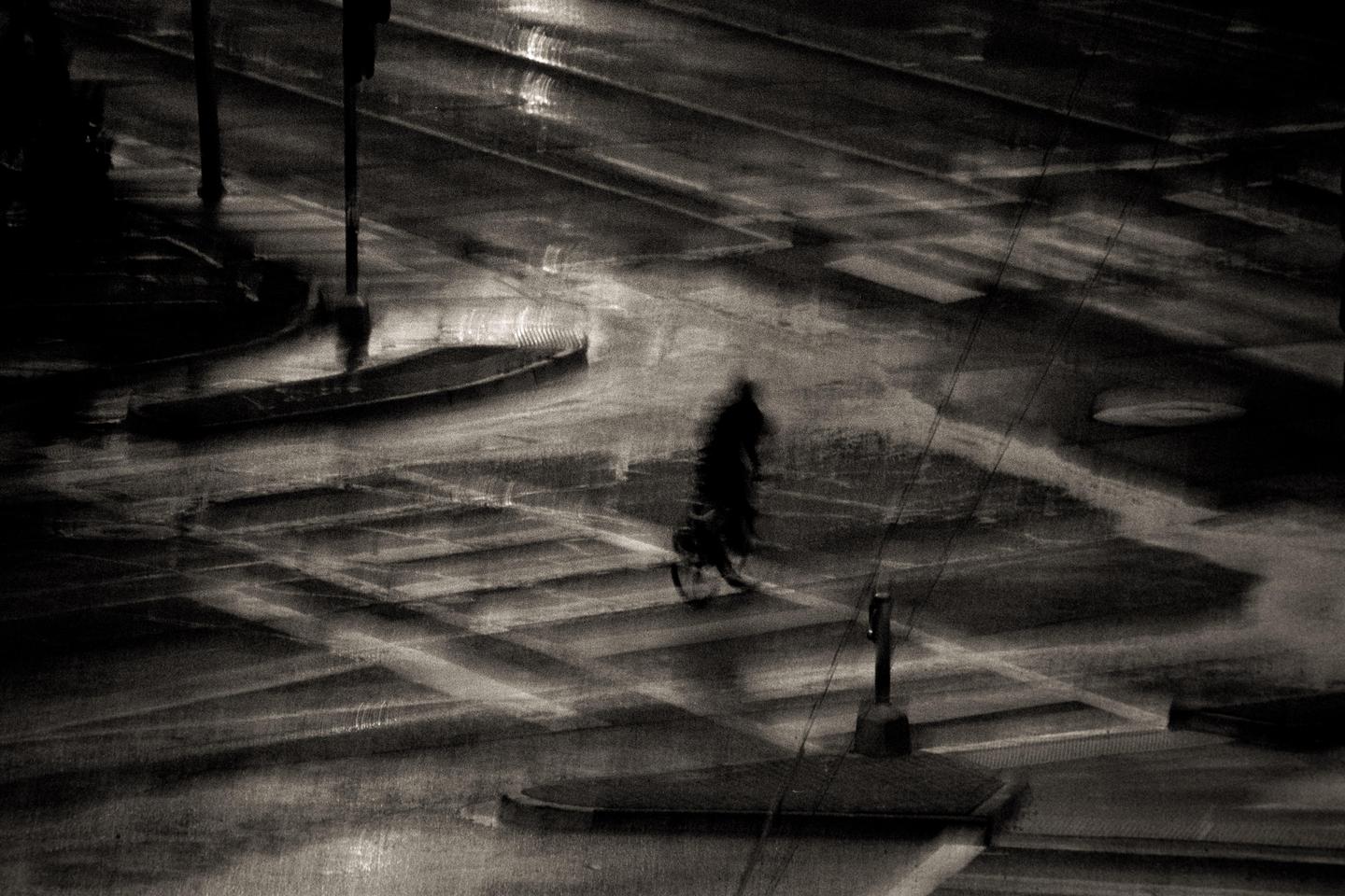 A blurry figure walks across a wet street at night, with reflections of light on the ground.