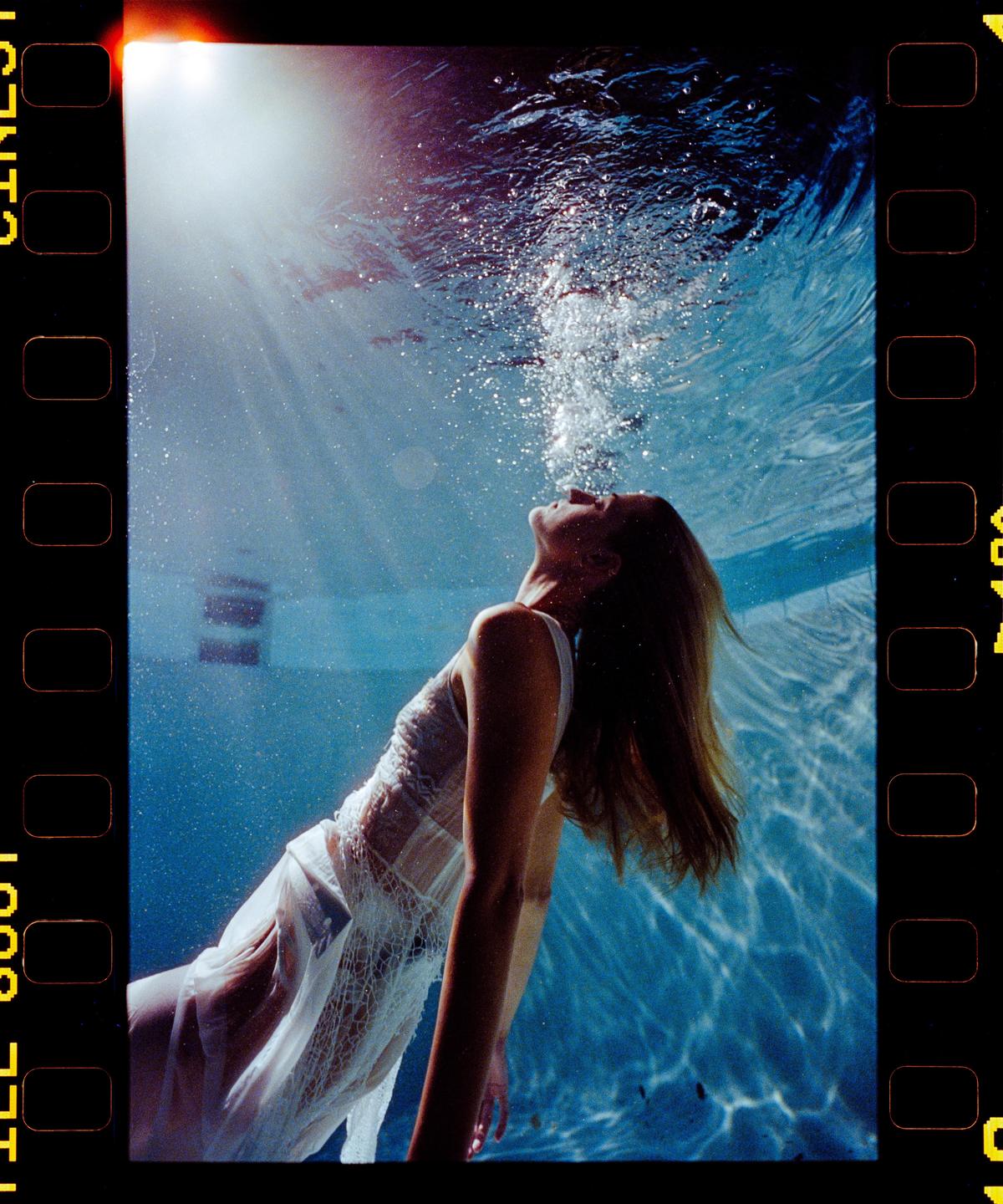A woman in a white dress is submerged in a swimming pool, looking up towards the surface as bubbles rise around her.