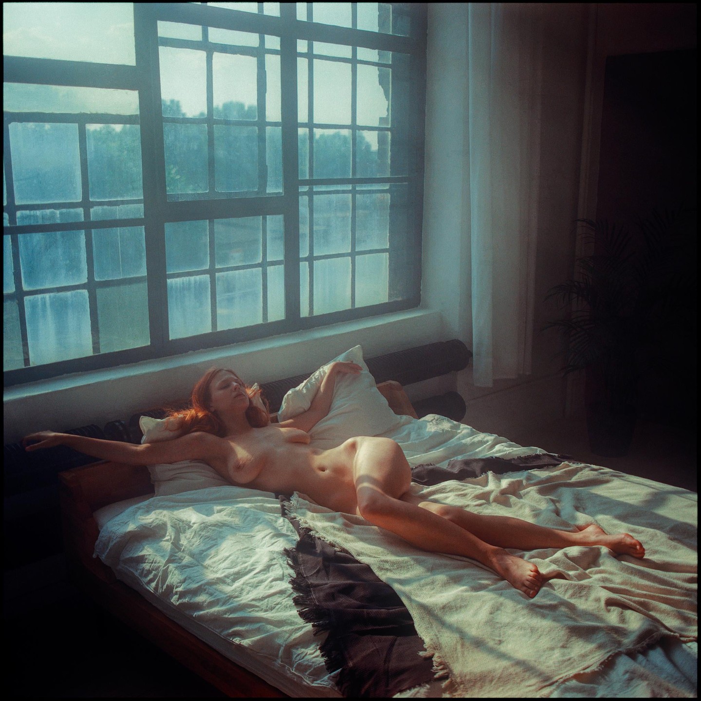 A nude woman with red hair lies on a bed next to a large window.