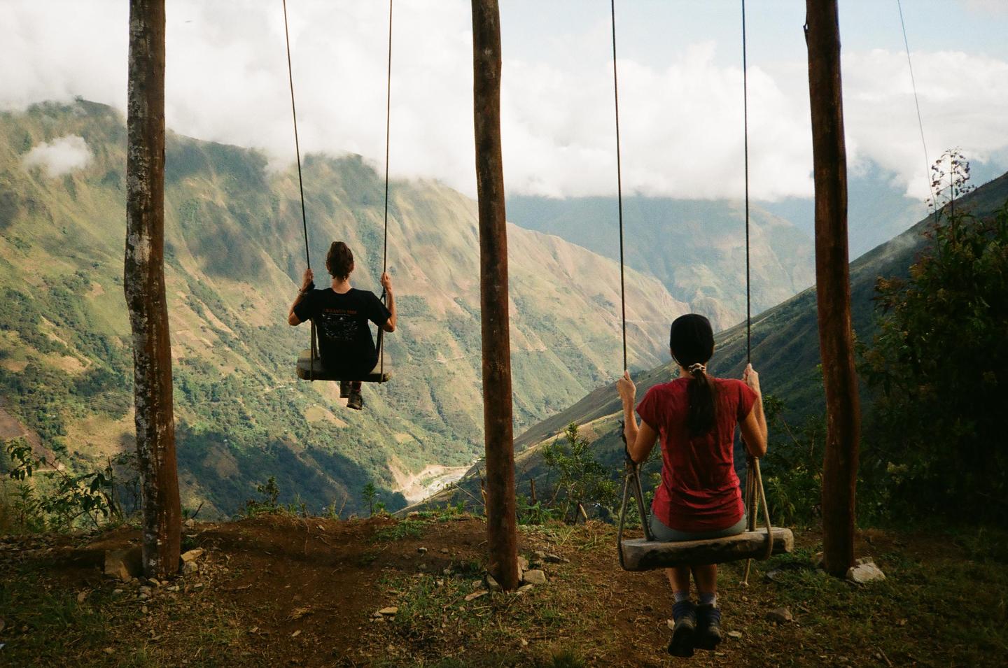 Two women sit on swings overlooking a valley with mountains and clouds.