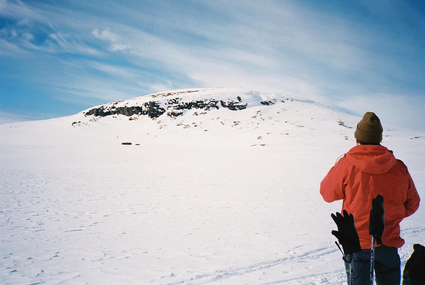 A person in a red jacket and a brown hat stands in a snowy landscape looking towards a snow-covered mountain.